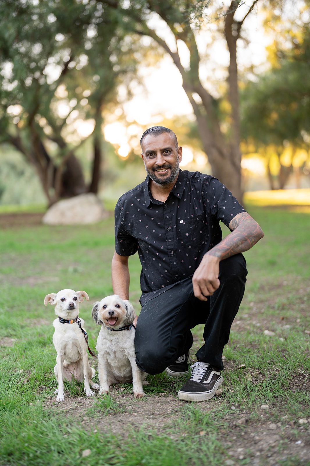 Dipan Desai, a professional family and pet photographer based in Los Angeles, kneeling in a park with two small white dogs during golden hour.